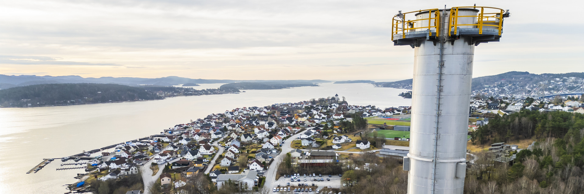 Utsikt över en Brevik i Norge med hus och en hamn längs vattnet, sedd från höjden av en vit industriskorsten till höger i bilden. Skorstenen har en gul säkerhetsplattform högst upp.