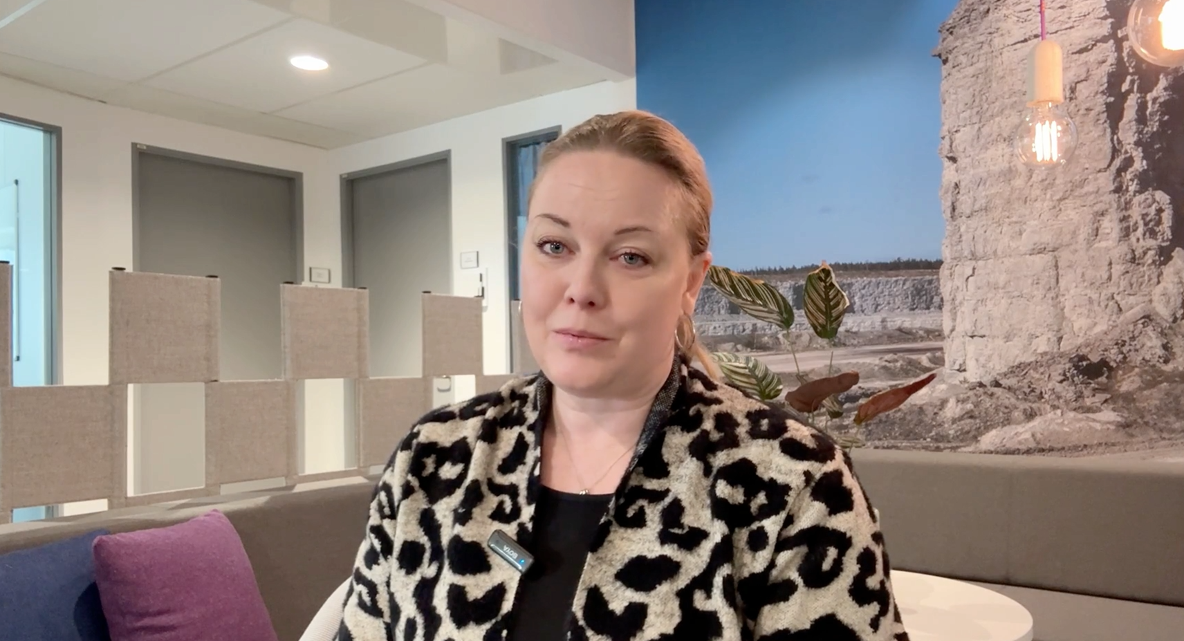 Person seated indoors on a modern office-style couch with a purple cushion, wearing a leopard-print jacket. The background features a lit-up wall with a coastal photo and minimalist partition panels.