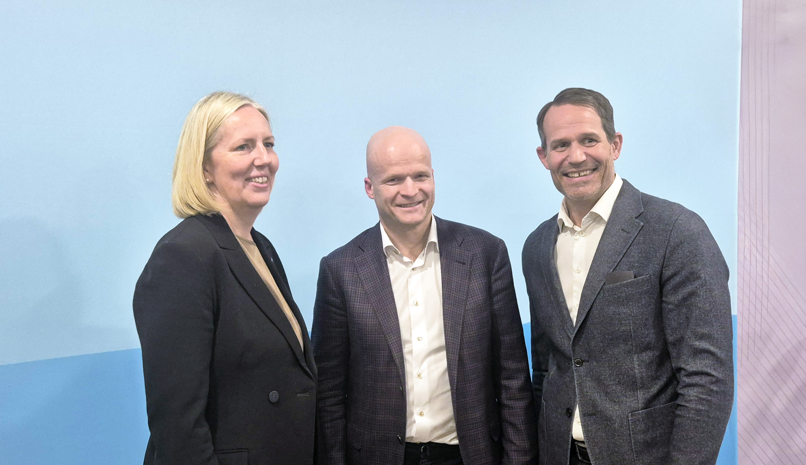 Tre personer står bredvid varandra framför en ljusblå bakgrund med en rosa ton i högra hörnet. De är klädda i formella kläder, inklusive kavajer och skjorta.