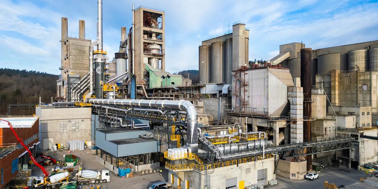 Industrial cement factory with large interconnected buildings, silos, and smokestacks under a blue sky. Machinery, vehicles, and equipment are visible in the foreground, with surrounding greenery in the distance.