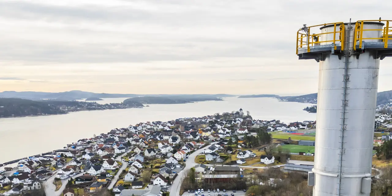 Utsikt over et kystområde med bolighus som strekker seg langs vannet til venstre. Til høyre står et høyt, hvitt tårn med en gul sikkerhetsplattform øverst. Skog og åser er synlige i bakgrunnen under en lys, overskyet himmel.