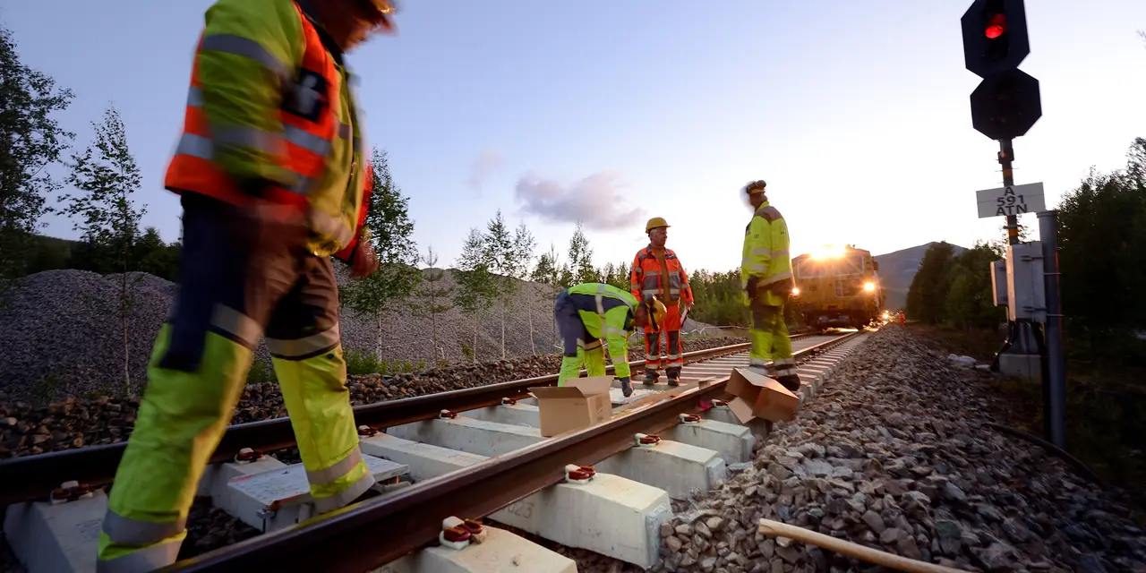 Arbetare i reflexkläder reparerar ett järnvägsspår i skymningen, med ett rött stoppljus till höger och skog i bakgrunden.