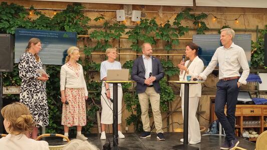 Fem personer står på en scen och deltar i en paneldiskussion med mikrofoner och en laptop.