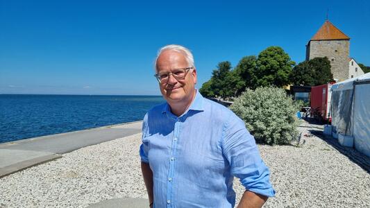 En person står vid en stenig strand med havet och en tornbyggnad i bakgrunden.